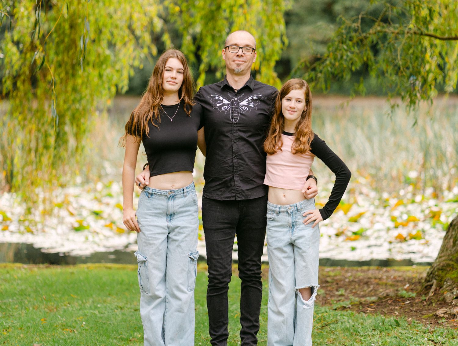 A man and two teenage girls pose together outdoors near a pond with willow trees in autumn.