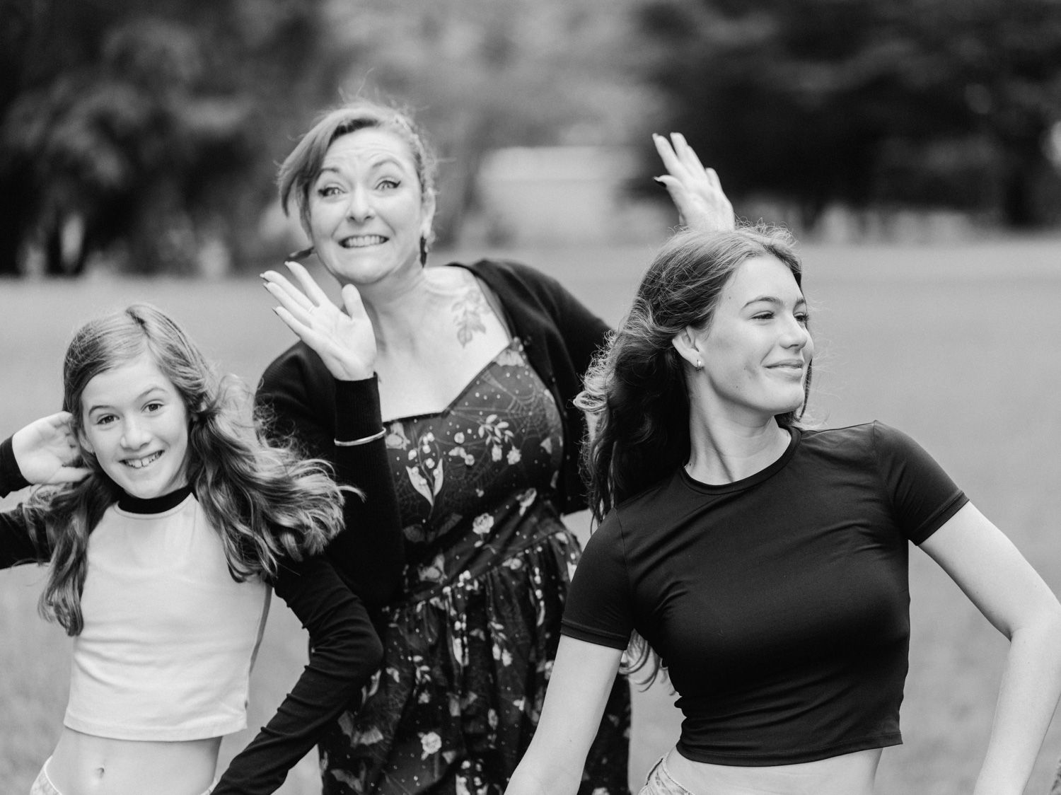 Three women smile and wave outdoors in a black and white photo taken in a park.