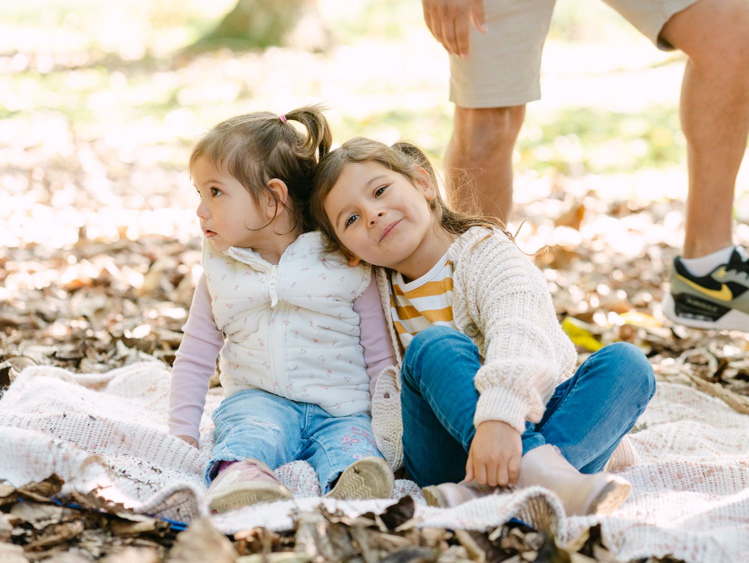 Two young girls sitting together on a blanket outdoors, leaning on each other among autumn leaves.