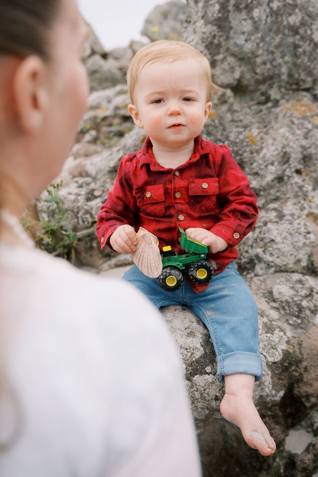 Toddler in red plaid shirt and jeans sits on rocks holding toy tractor while mother offers a shell.