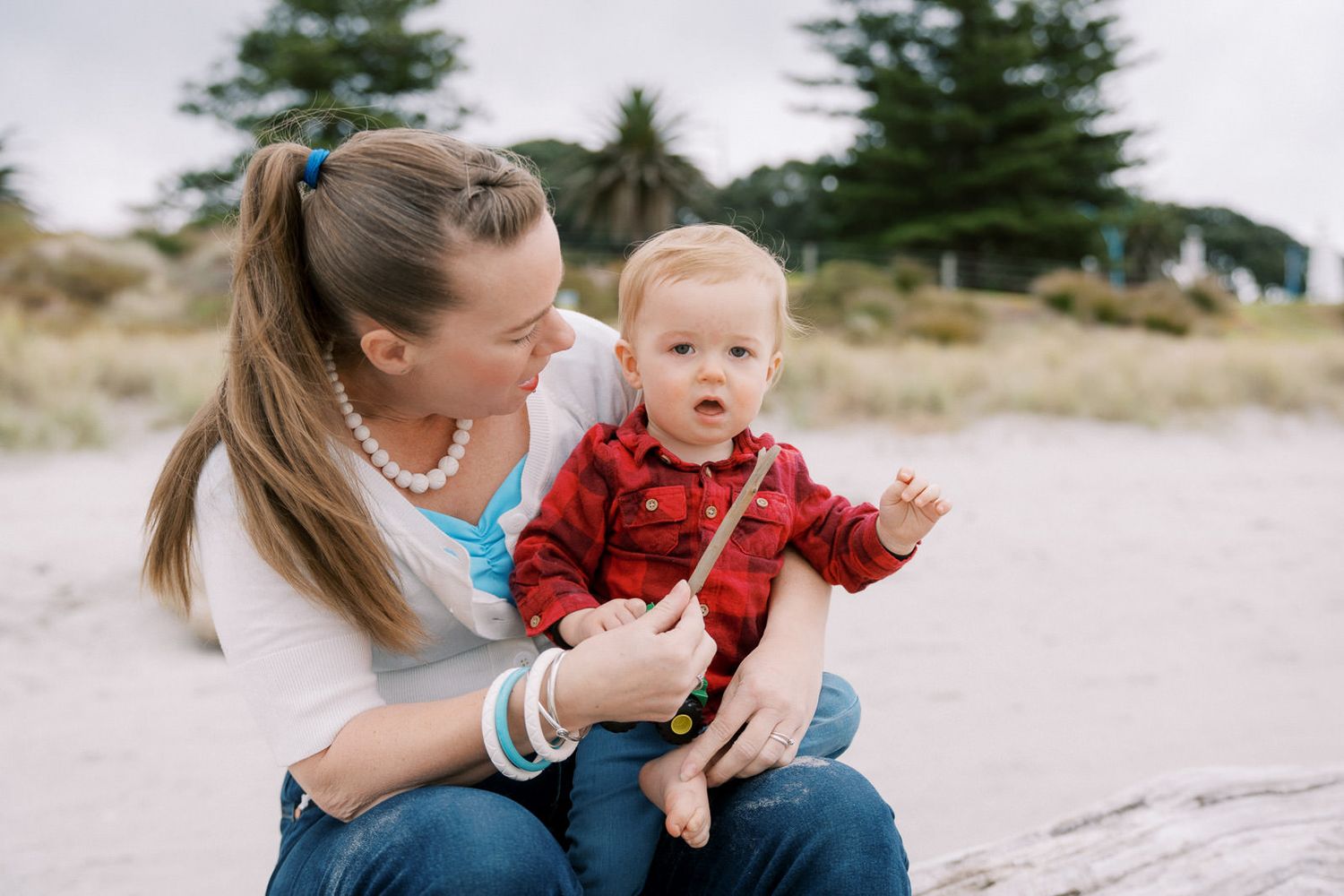 A mother sits on a sandy beach holding her baby boy in a red plaid shirt, both enjoying a sunny outdoor moment.