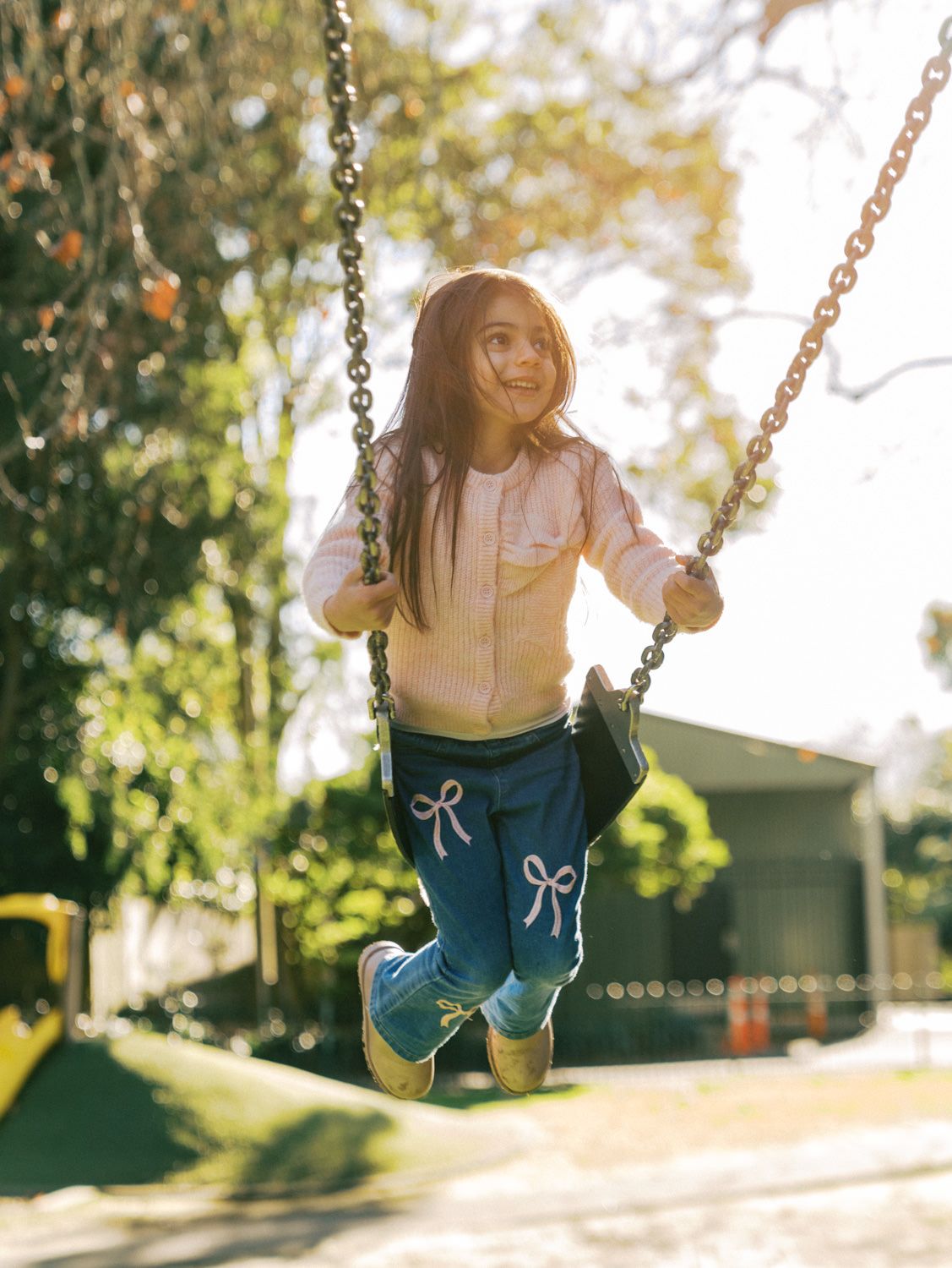Young girl with long hair smiling on a chain swing outdoors, wearing pink jacket and blue bow pants.
