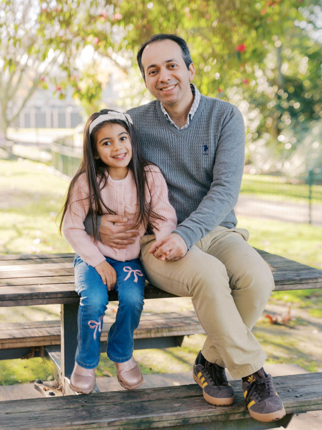 A man and young girl smile together on a wooden bench in a sunny park setting.
