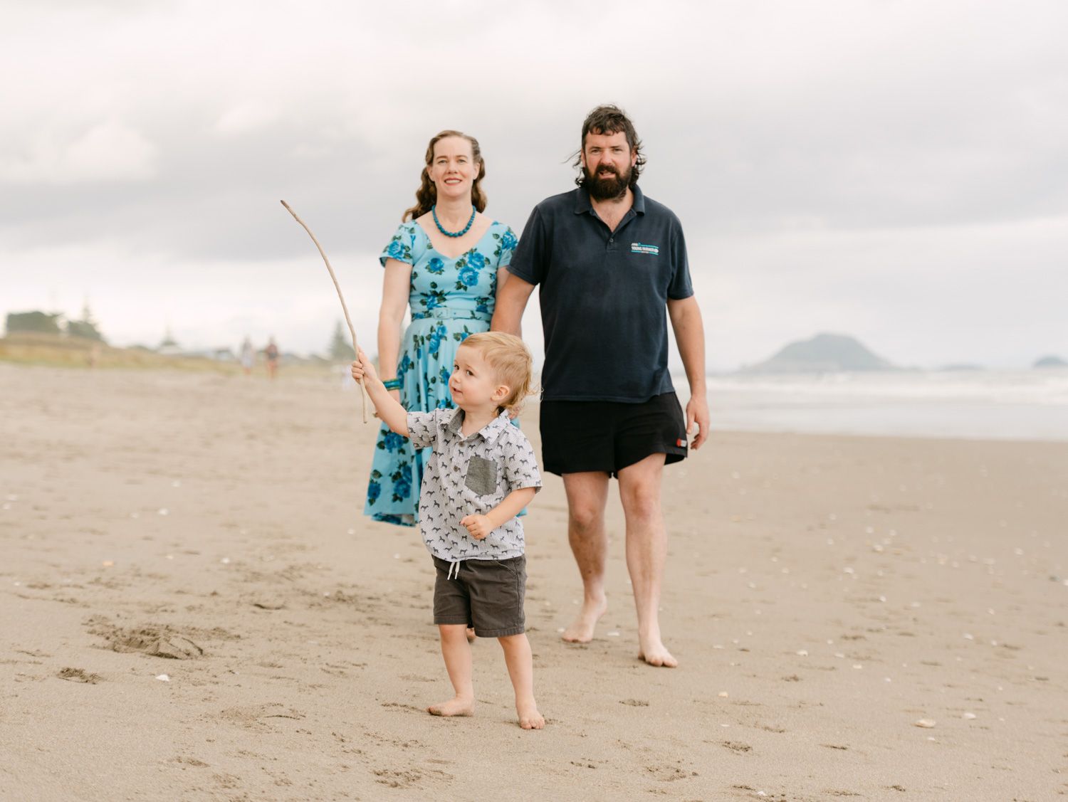Family of three walking on a sandy beach, woman in floral dress, bearded man, and young child holding a stick.