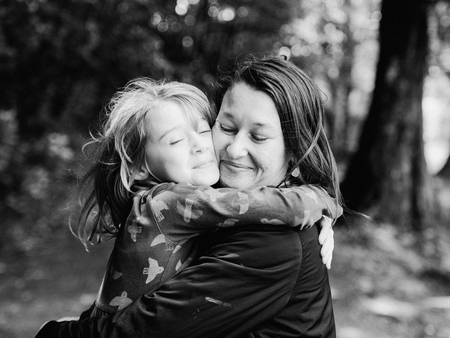 A black and white photo of a child tenderly hugging a smiling woman among trees in a heartfelt outdoor moment.