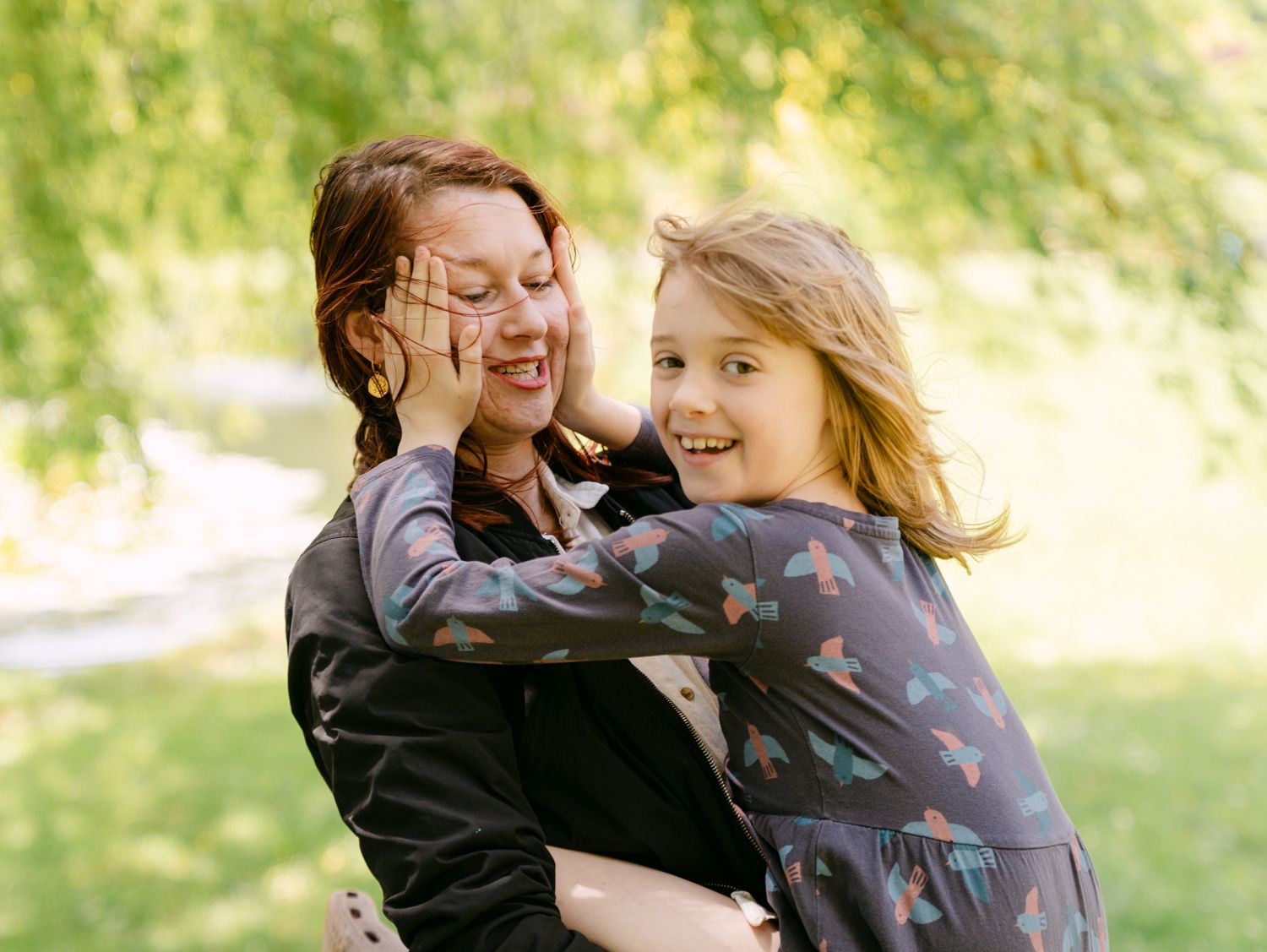 A mother and daughter share a joyful embrace outdoors, with the girl playfully covering her mom's face while smiling.