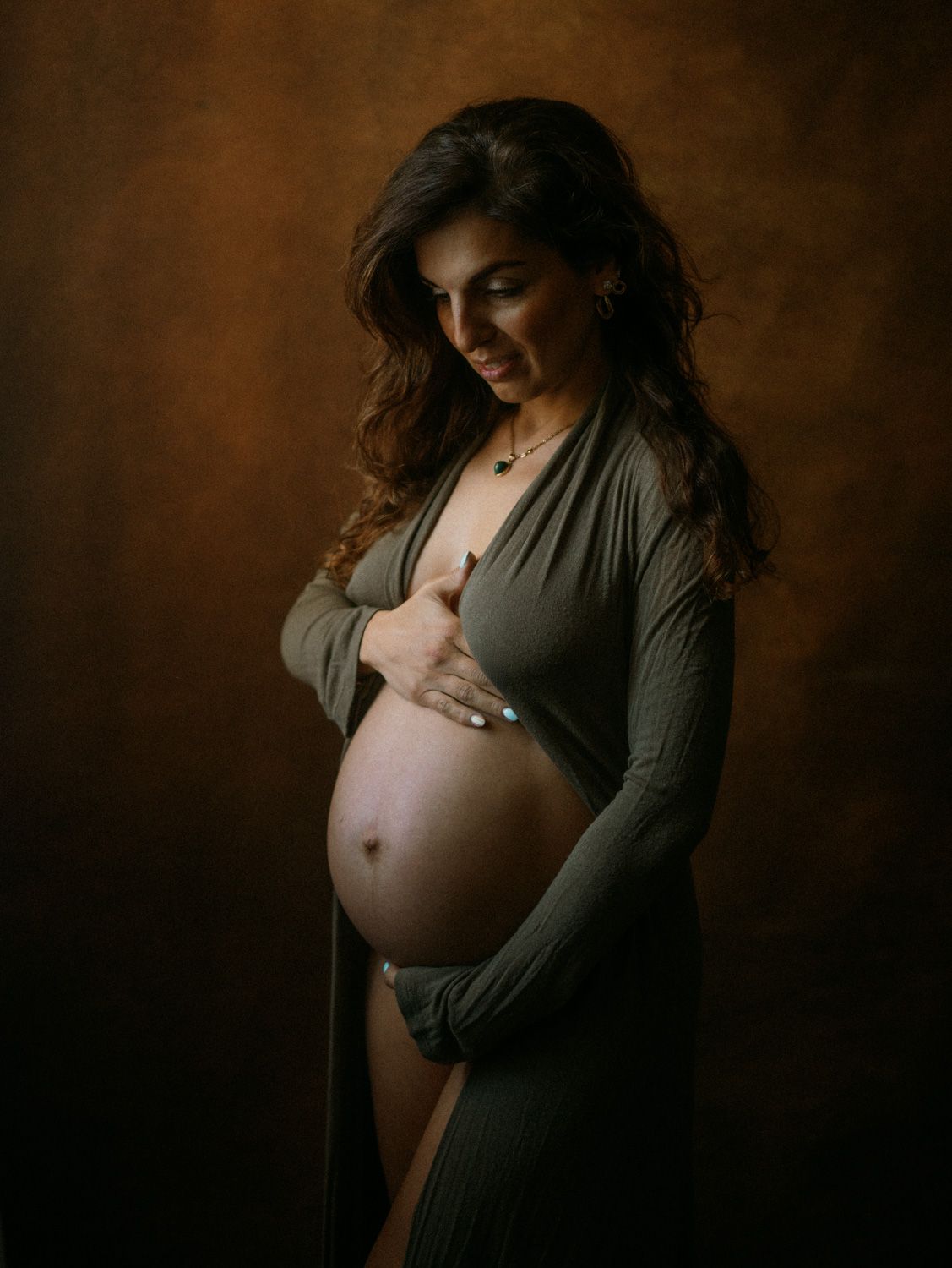 Pregnant woman in olive green wrap dress cradling her baby bump against a warm brown studio background.