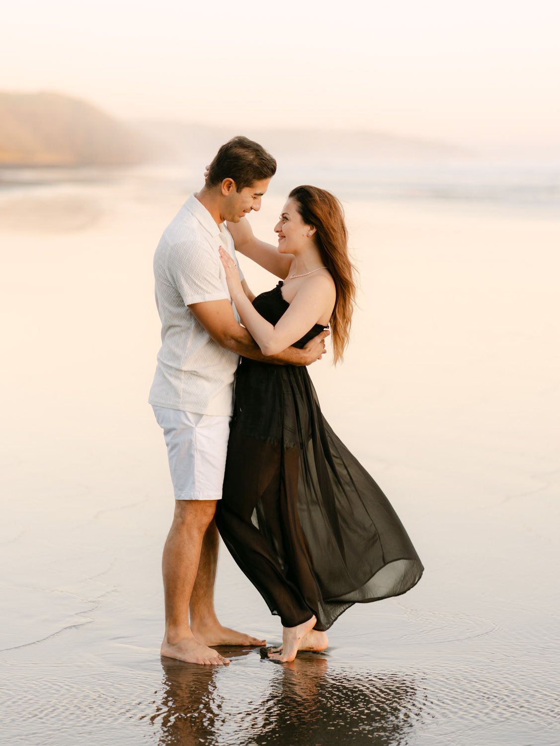 Romantic couple embracing on a misty beach at sunset, woman in flowing black skirt.