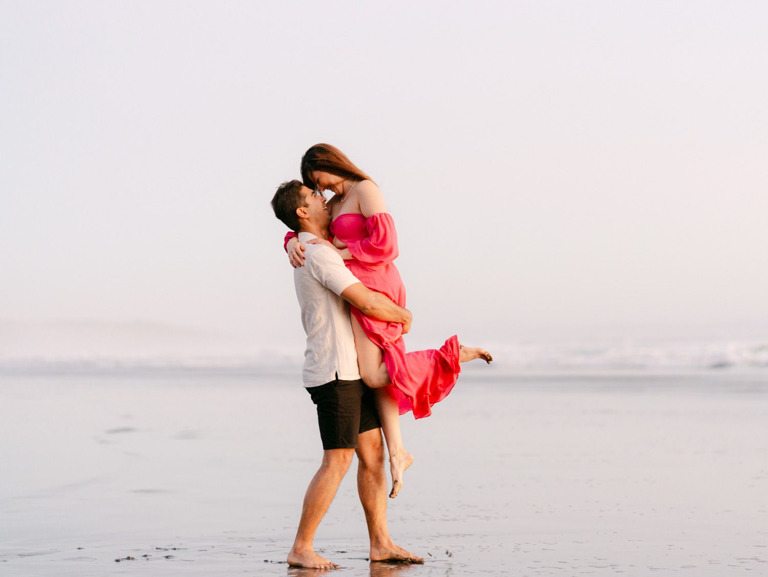 A romantic couple sharing a kiss on a beach, man lifting woman in pink dress near ocean waves.