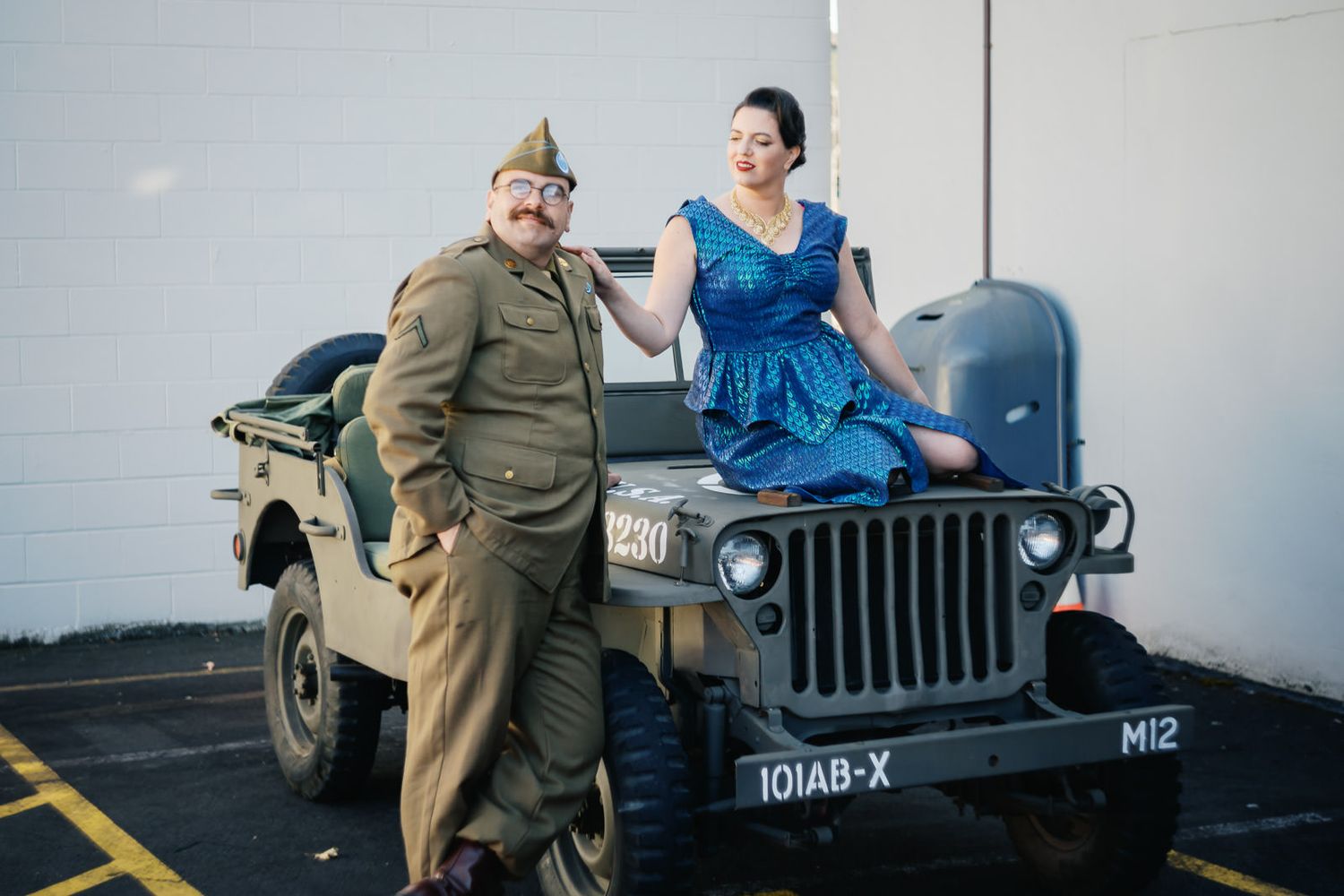 Man in WWII military uniform and woman in blue dress posing beside a vintage olive green military jeep.