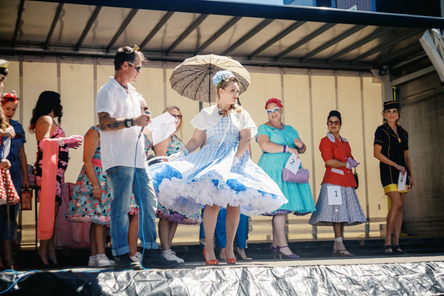 Women in vintage 1950s dresses dance on stage at a retro fashion show, one twirling a parasol outdoors.