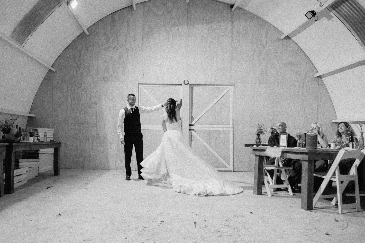 Black and white photo of bride and groom sharing first dance in rustic barn venue with wooden arch ceiling.