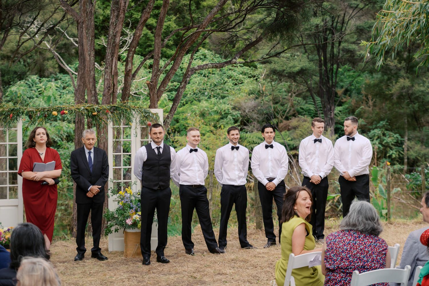 Outdoor wedding ceremony with groom and groomsmen in black bow ties standing before an officiant in a lush garden setting.