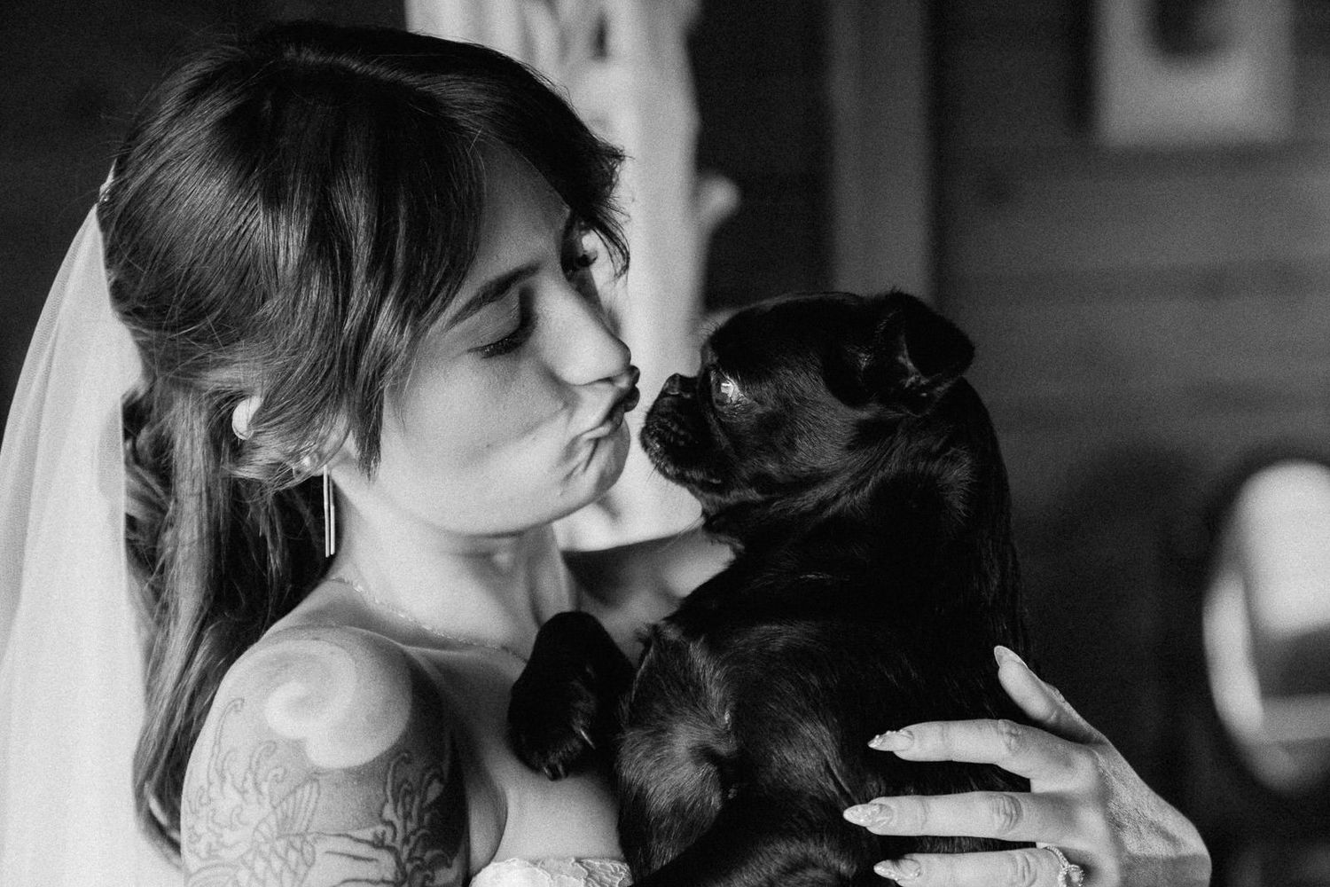 Black and white photo of a tattooed bride in veil kissing her small black dog on her wedding day.