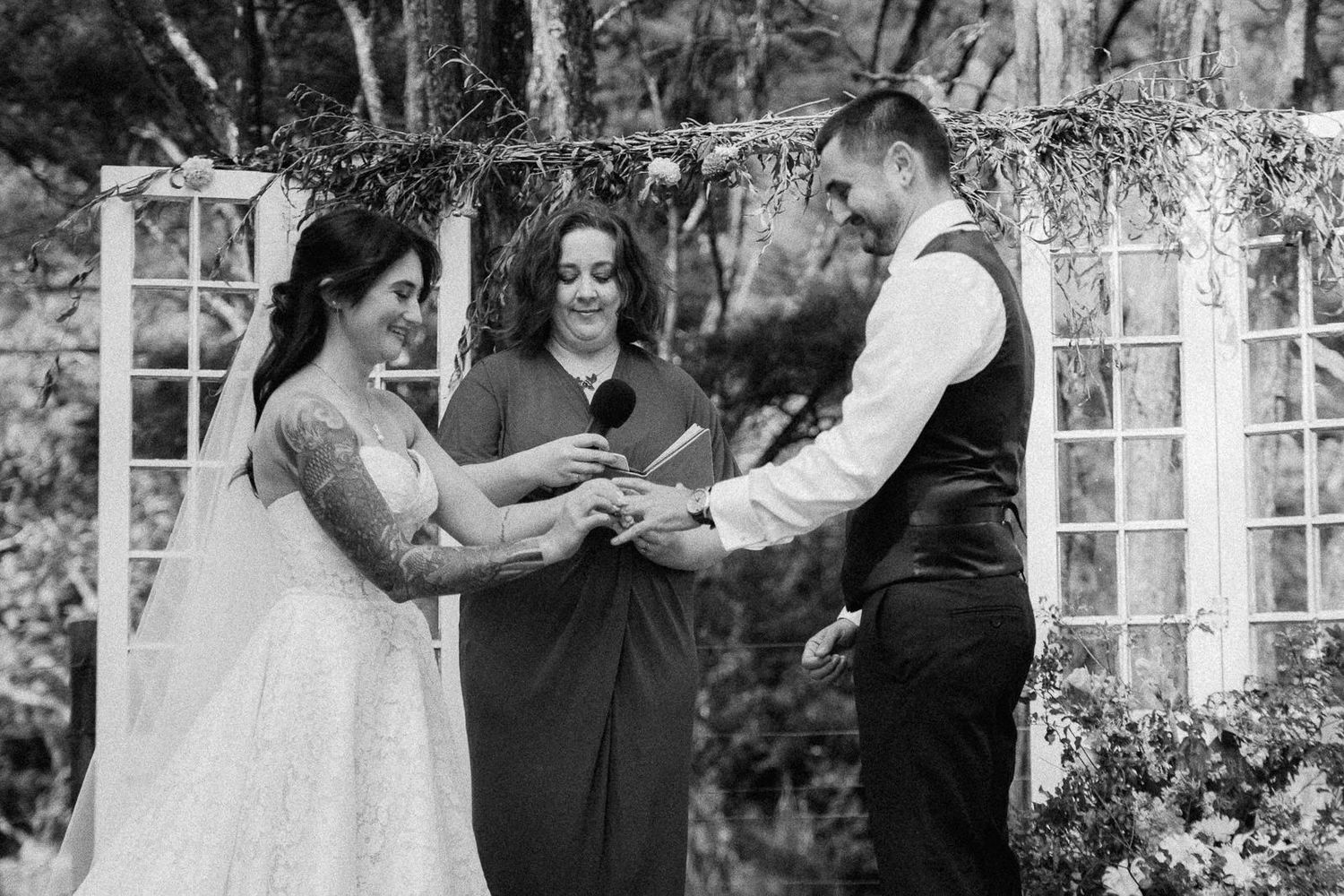 Black and white photo of bride placing ring on groom's finger during outdoor wedding ceremony with officiant.