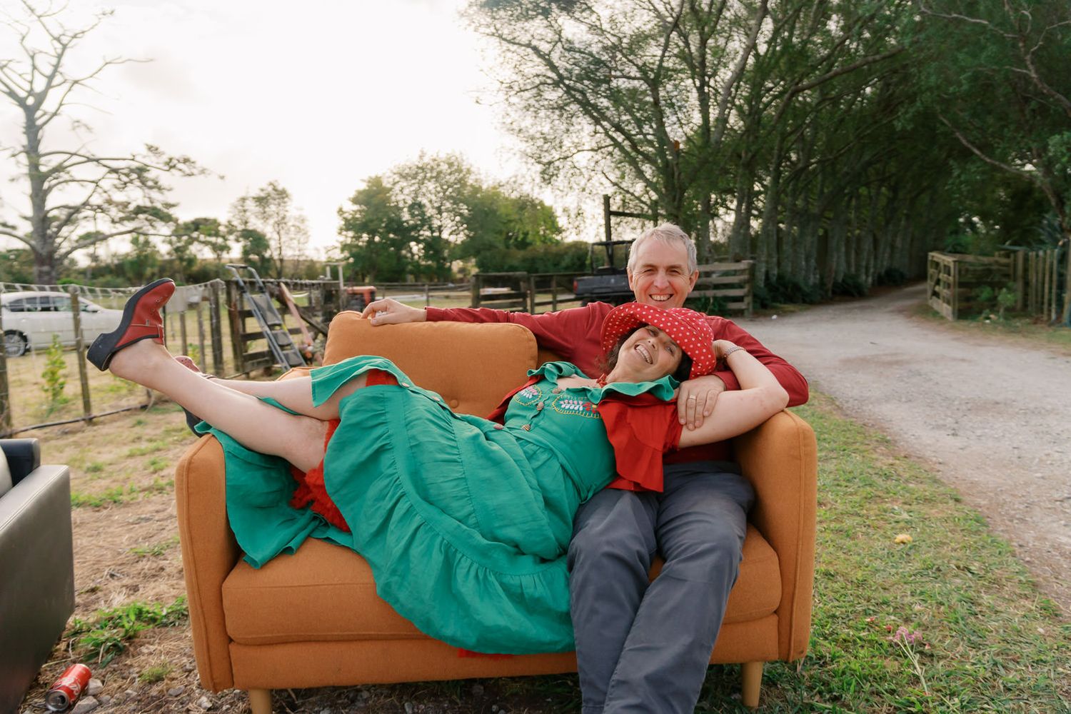 Two people laughing and relaxing on an orange sofa outdoors in a garden setting, wearing green and red outfits.