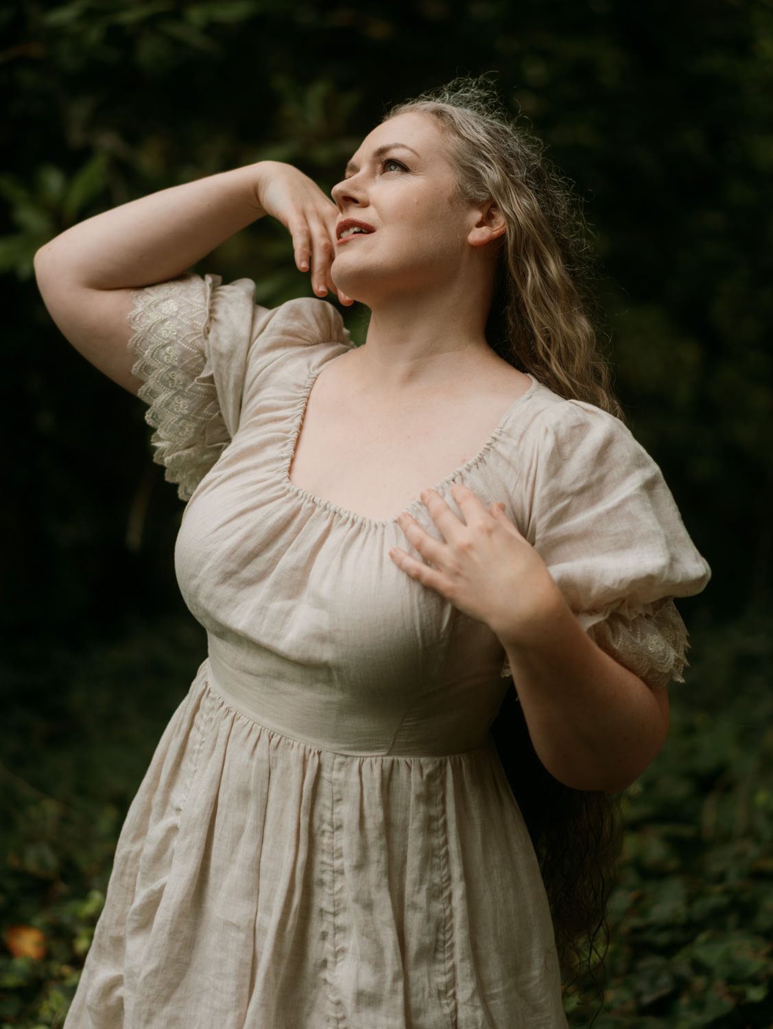 Woman in vintage cream lace dress posing outdoors among lush green foliage.