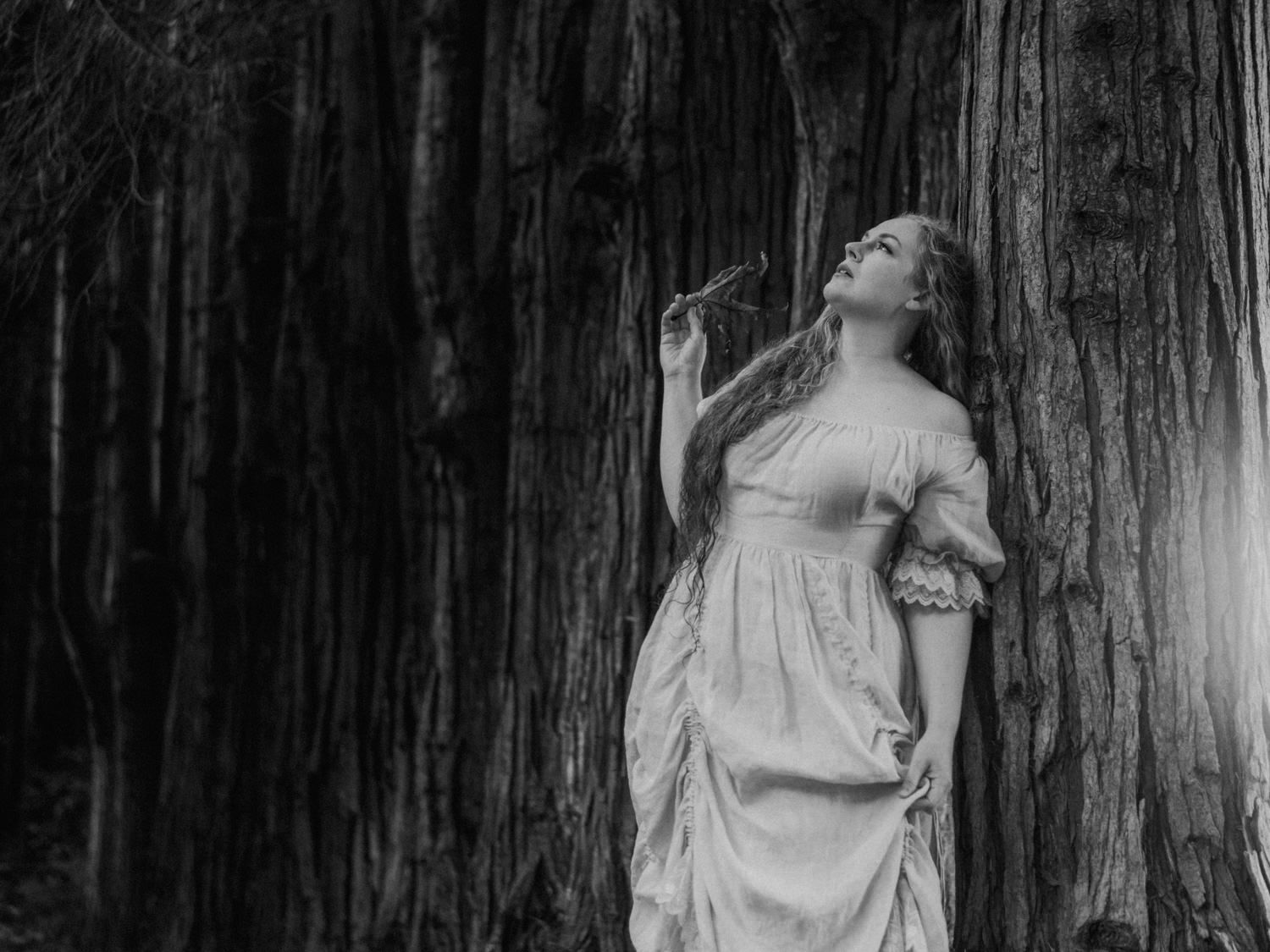 Black and white photo of a woman in a white dress leaning against a large tree in a forest.