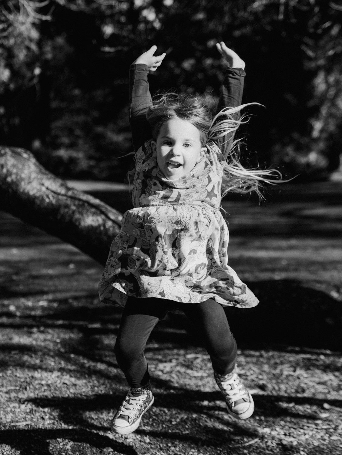 A young girl leaps with arms raised and mouth open in a playful black-and-white outdoor park photo.