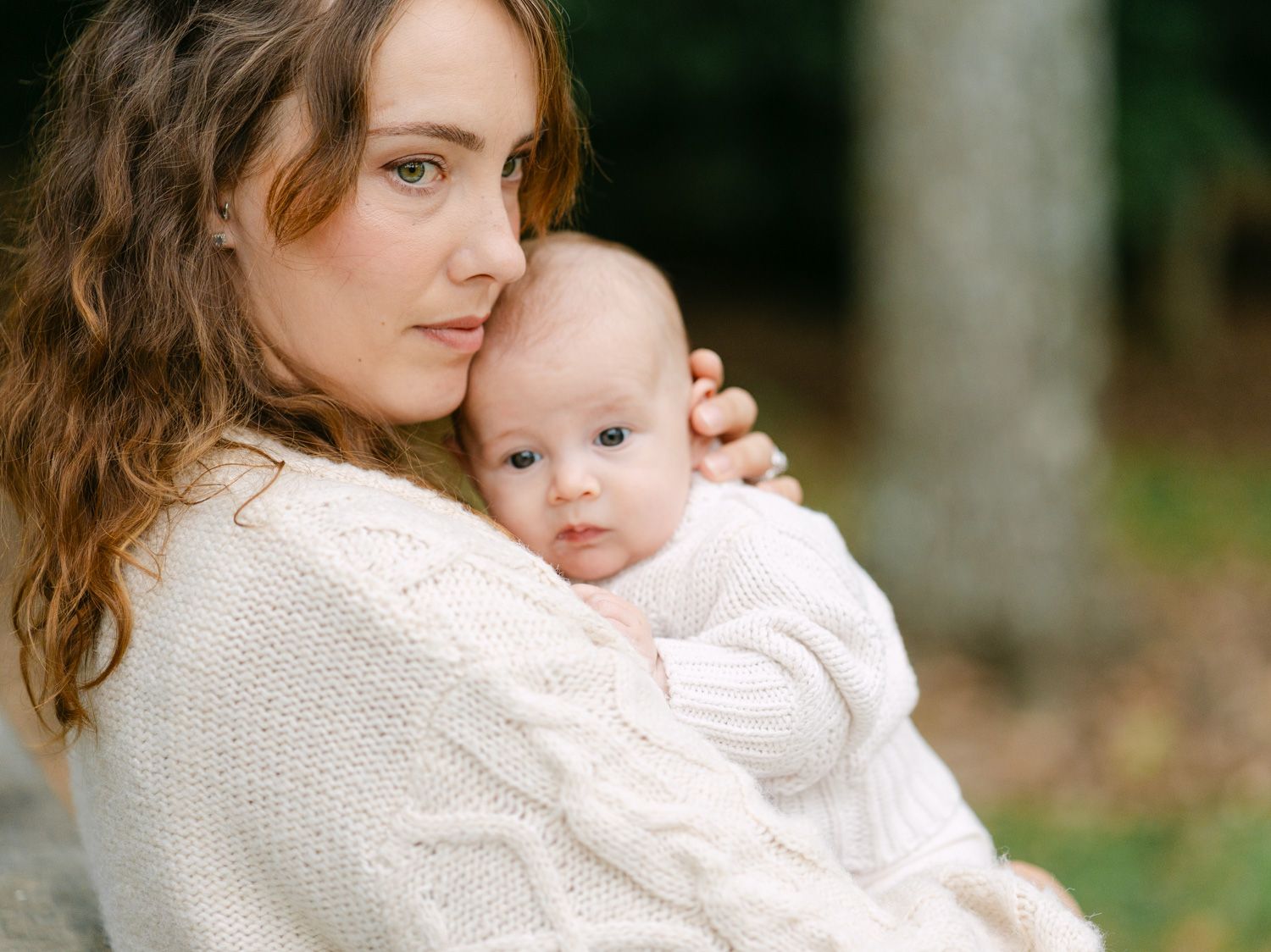 A young mother in a white cable-knit sweater tenderly holds her infant baby outdoors in a serene natural setting.