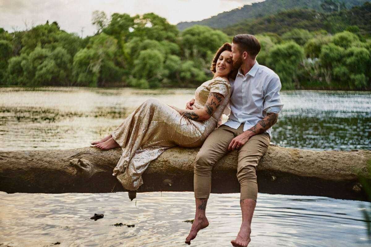 Romantic couple sitting on a log by a serene lake, surrounded by lush green trees in a peaceful outdoor setting.