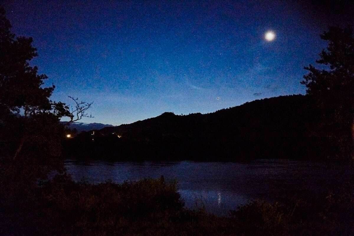 Moonlit night sky over a serene mountain lake with dark silhouetted hills and trees reflecting on calm water.
