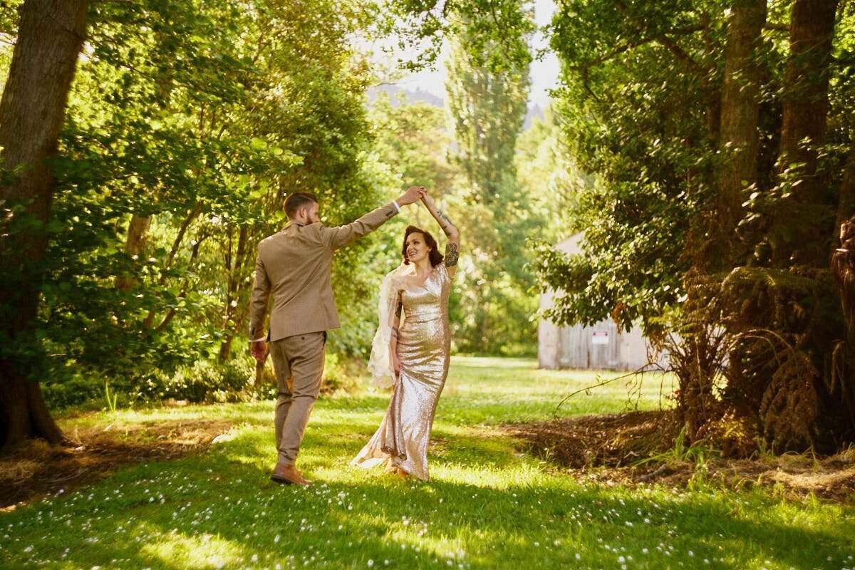 Couple dancing outdoors in lush green garden, bride in gold dress, groom in tan suit, spinning under raised hands.