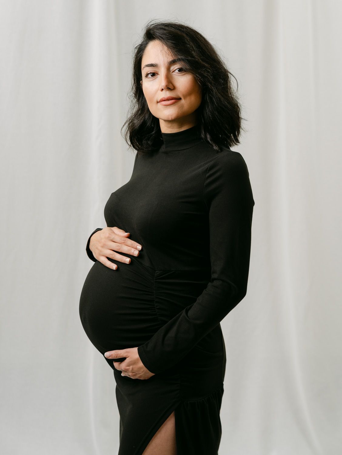 Pregnant woman in black turtleneck dress cradling baby bump, maternity photo shoot on white background.