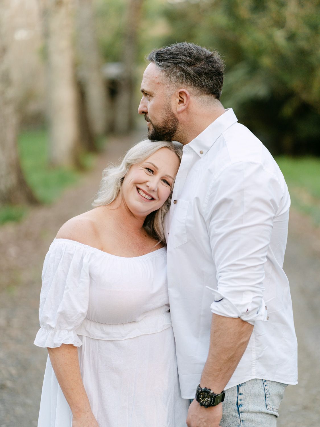 A smiling couple in white outfits share a tender moment on a tree-lined path, the man kissing her head.