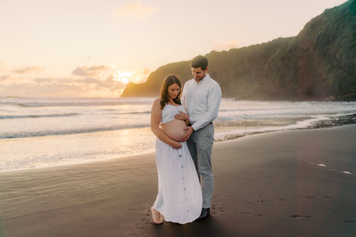 Expectant couple looking down at baby bump on black sand beach during golden hour sunset.