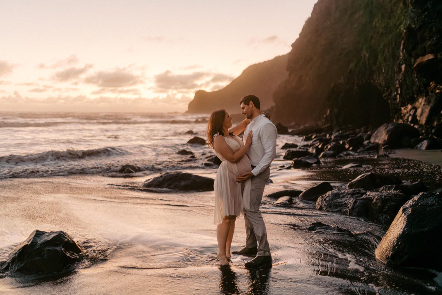 Couple embracing on a rocky beach at golden sunset with ocean waves and dramatic cliffs in background.