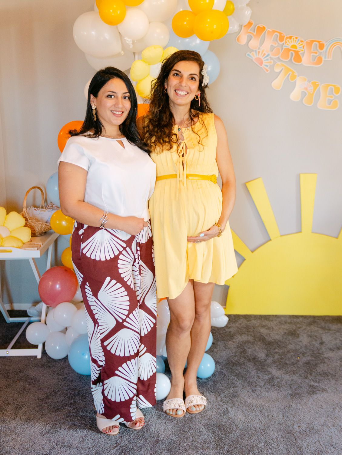 Two women pose at a sunny baby shower with yellow balloons and sun decorations, one wearing a yellow dress showing a baby bump.