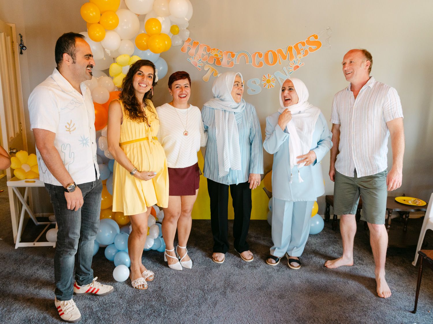 Group of six people smiling at a baby shower with yellow and white balloon decorations and a welcome baby banner.