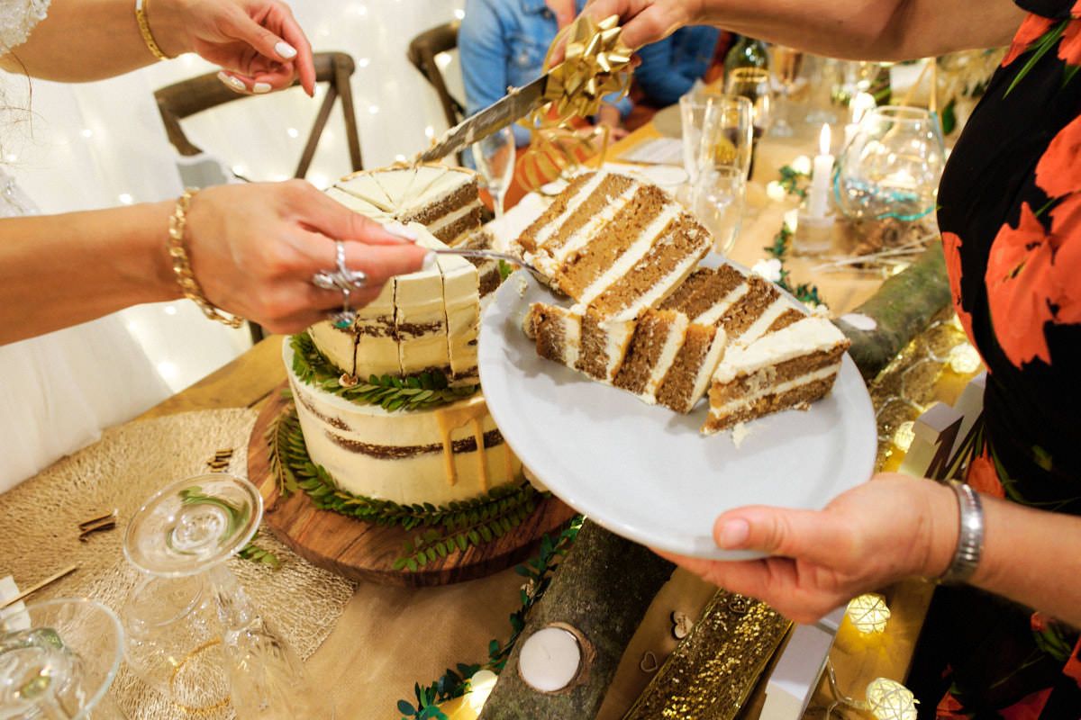 Guests cutting and serving slices of a layered naked wedding cake on a wooden board at a reception.