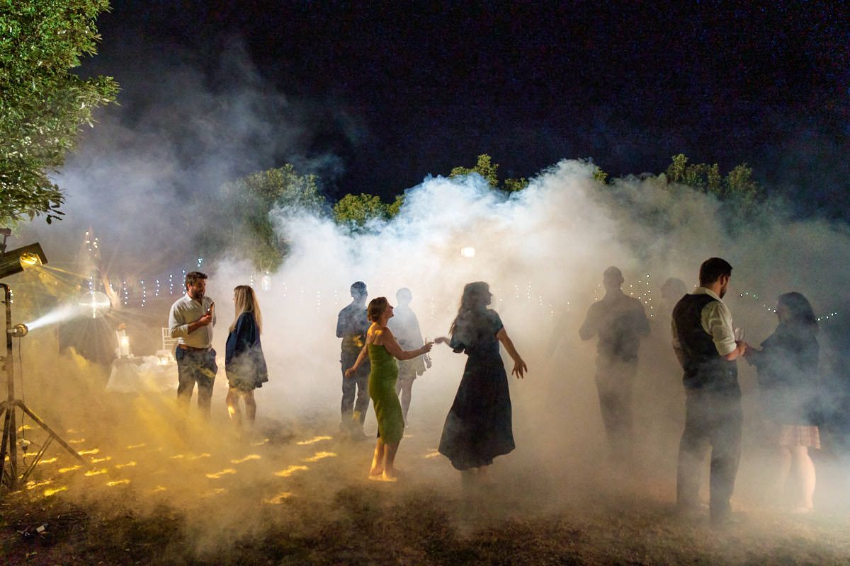 People dancing outdoors at night surrounded by dramatic smoke effects and warm golden lighting at an evening event.