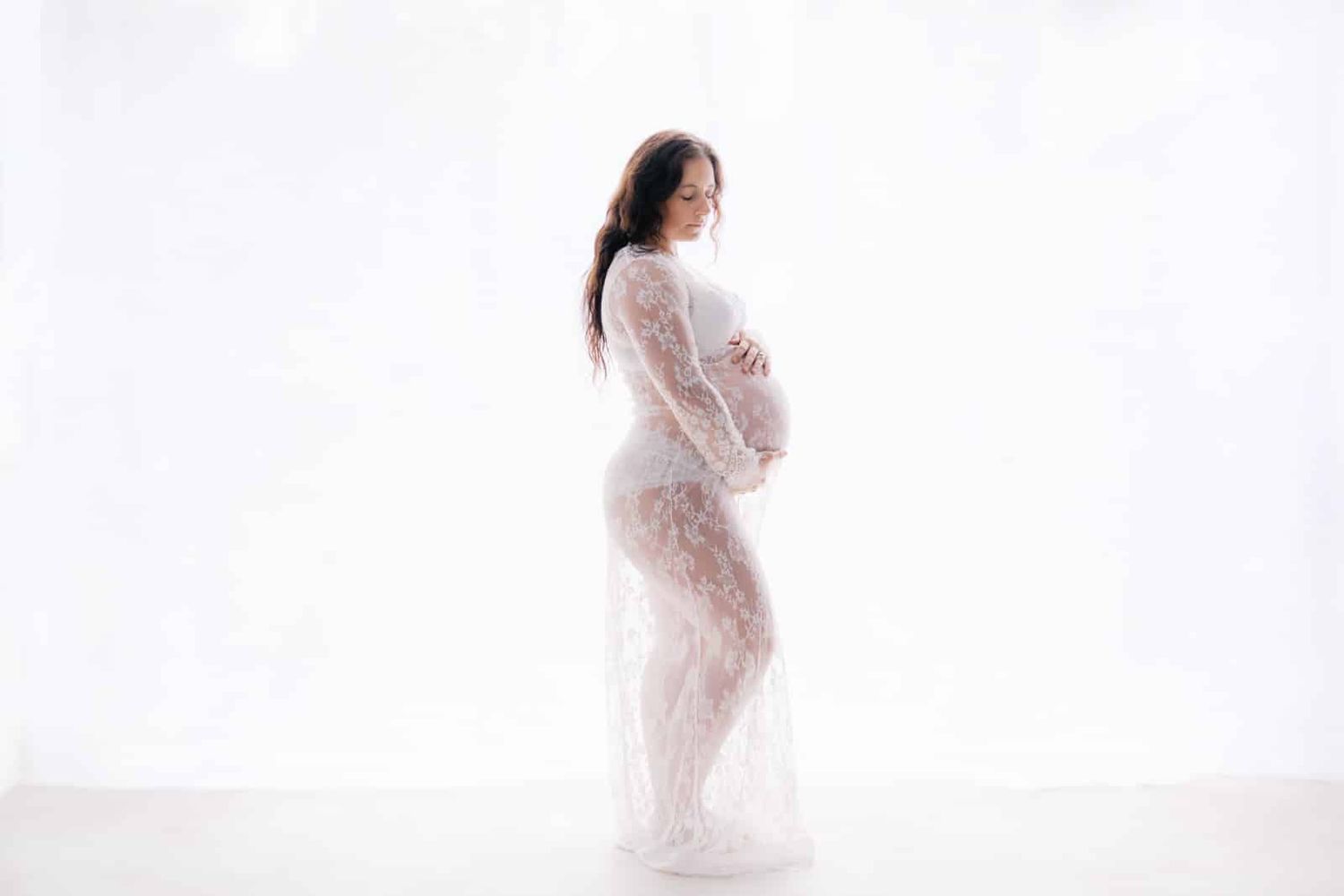 Pregnant woman in white lace maternity gown cradling baby bump against bright white studio background.
