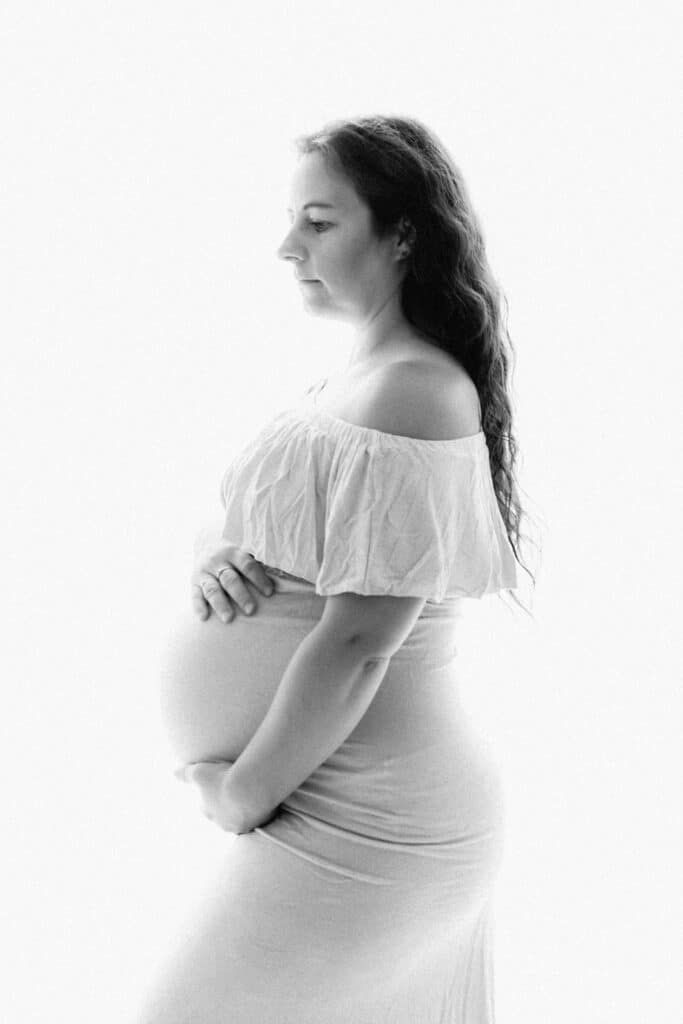 Black and white maternity photo of a pregnant woman in a white off-shoulder dress cradling her baby bump.