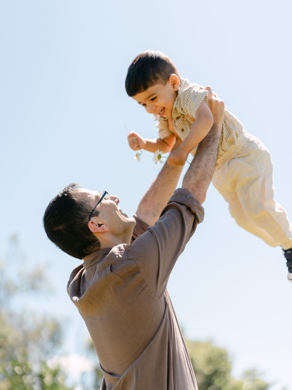 Parent joyfully lifting smiling toddler high in the air outdoors on a sunny day with blue sky background.