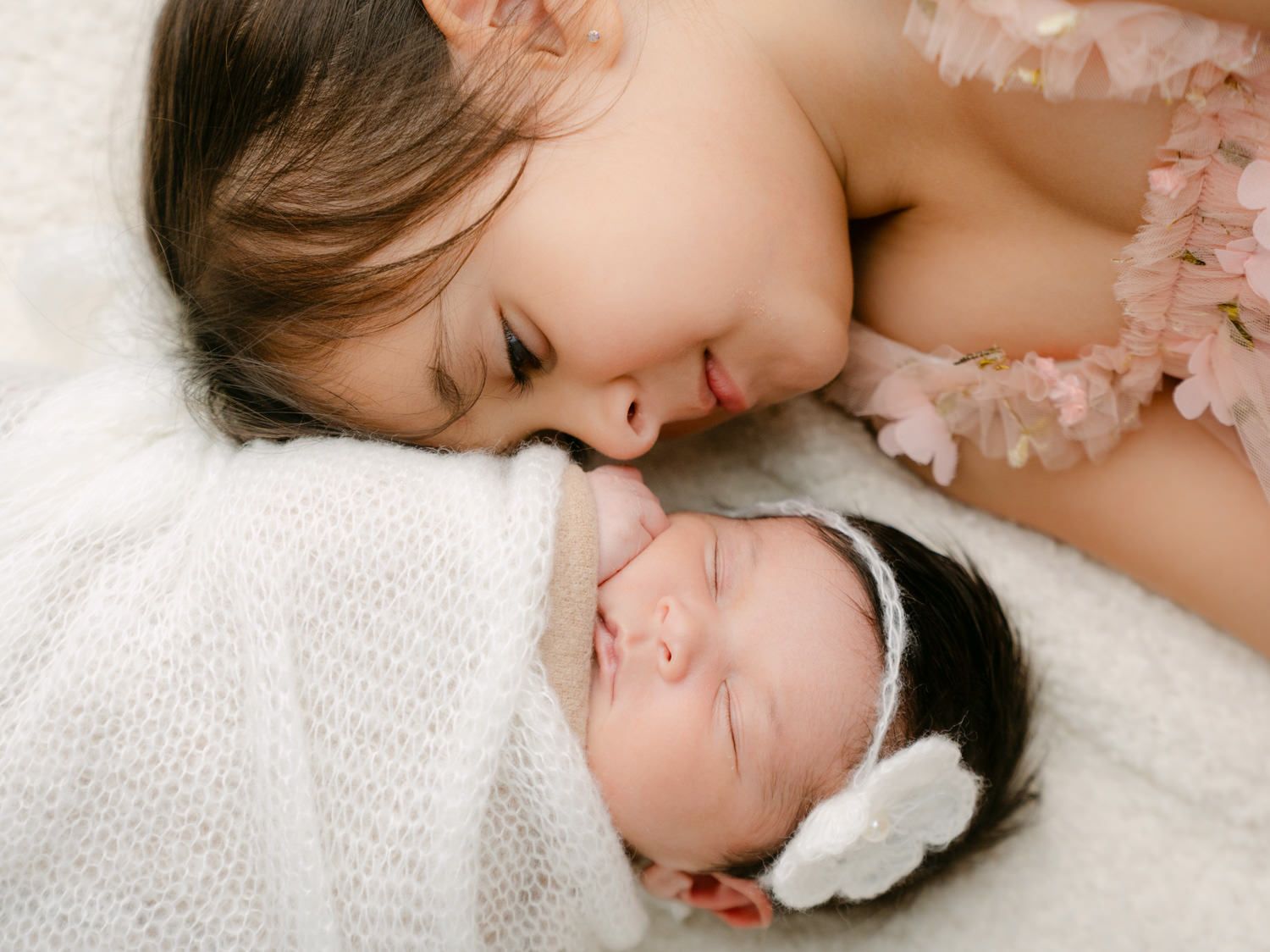 A young girl lovingly gazes at a sleeping newborn baby wearing a white bow headband.