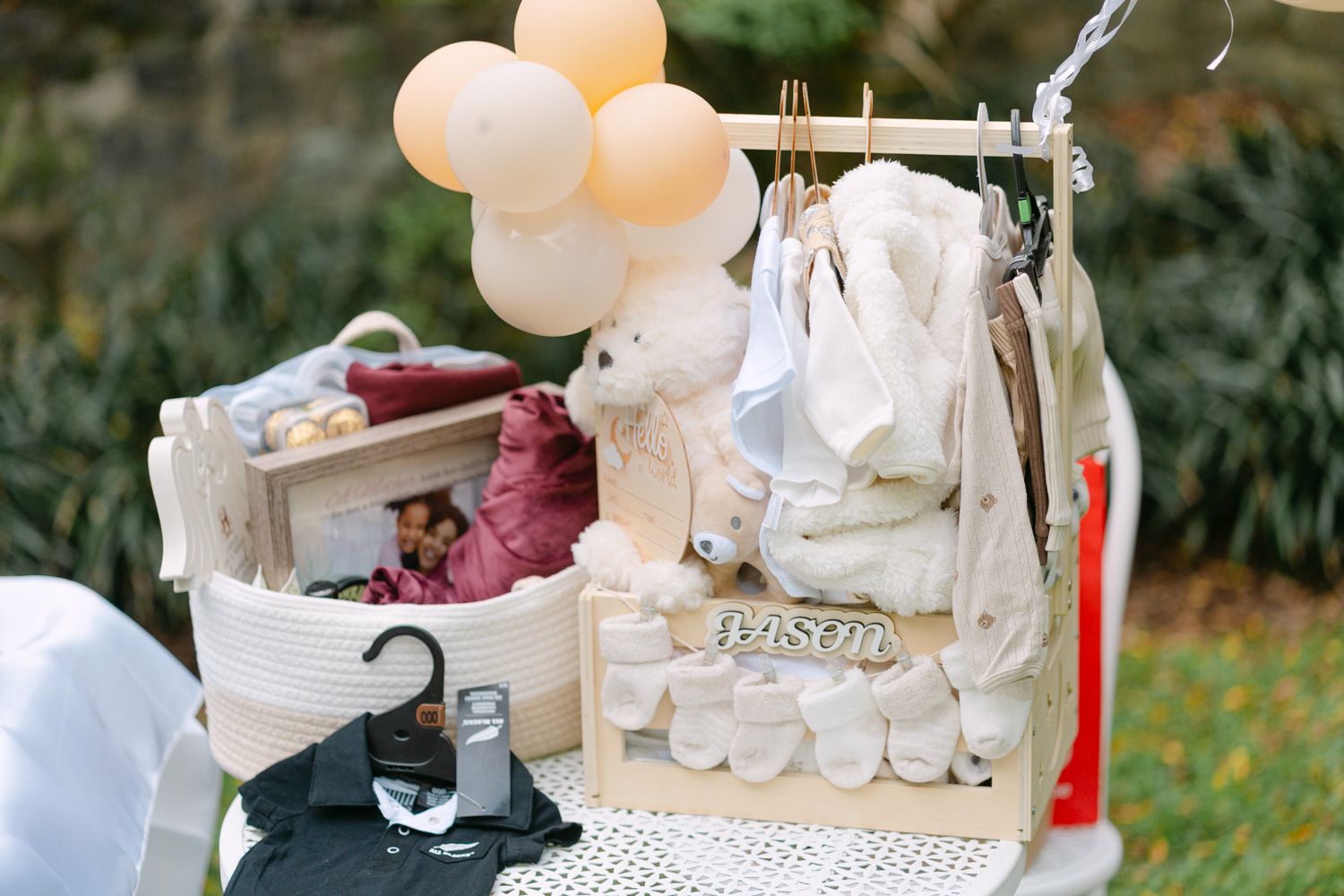 Baby shower gift display with wooden crates, stuffed bear, pastel balloons, and hanging infant clothes on a rack outdoors.