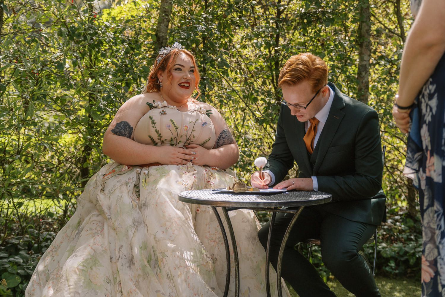 Bride in floral gown laughs as groom in navy suit signs wedding documents at outdoor ceremony table.