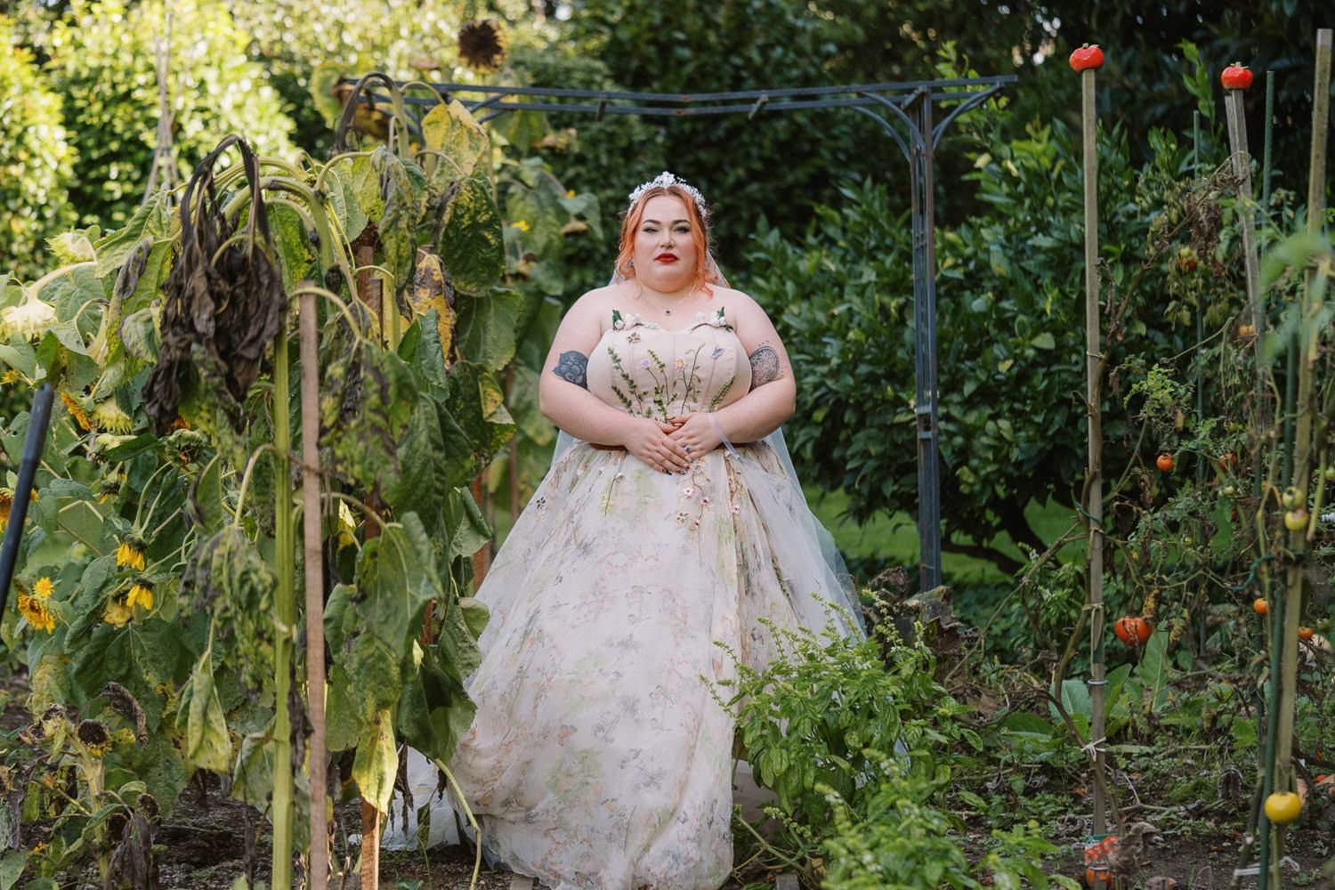 Woman in floral ballgown wedding dress standing in lush garden with greenery and tomato plants.