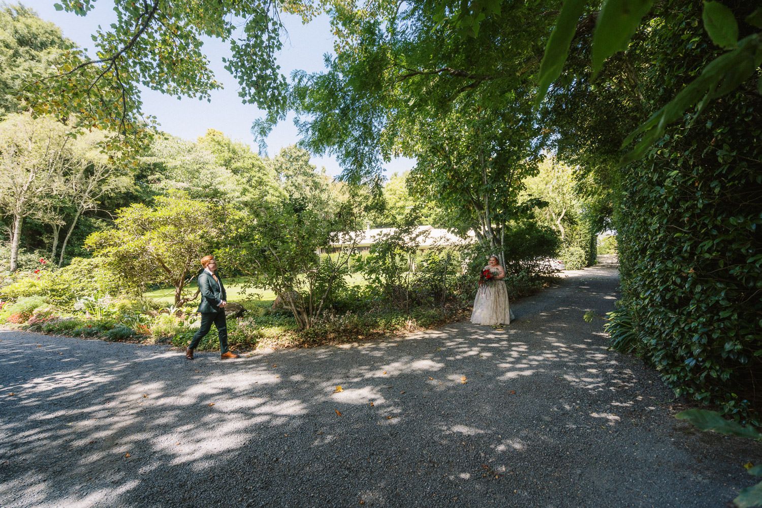Person walking along a scenic garden path surrounded by lush green trees and shrubs on a sunny day.