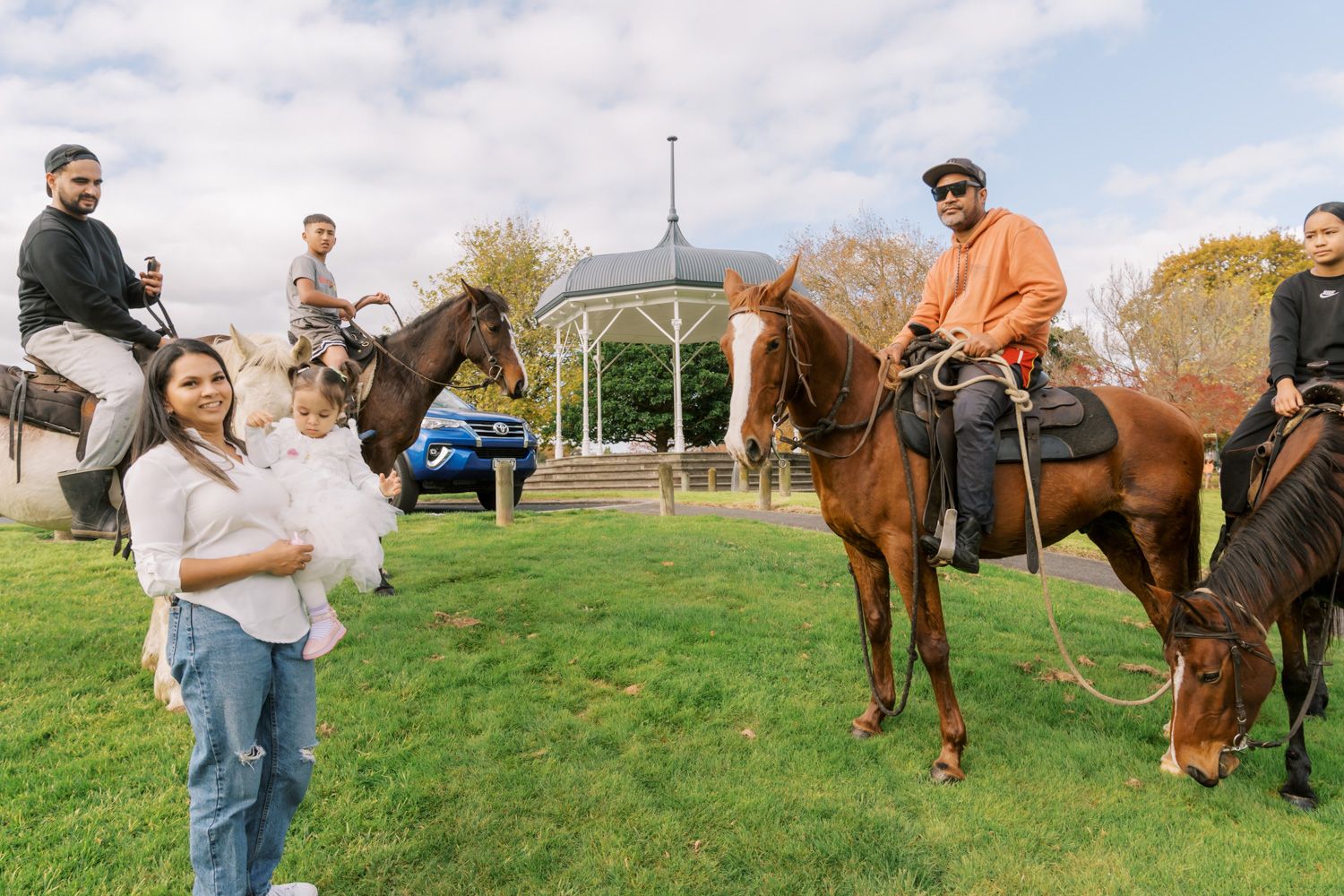 Group of people enjoying horseback riding in a scenic park with a gazebo on a cloudy day.