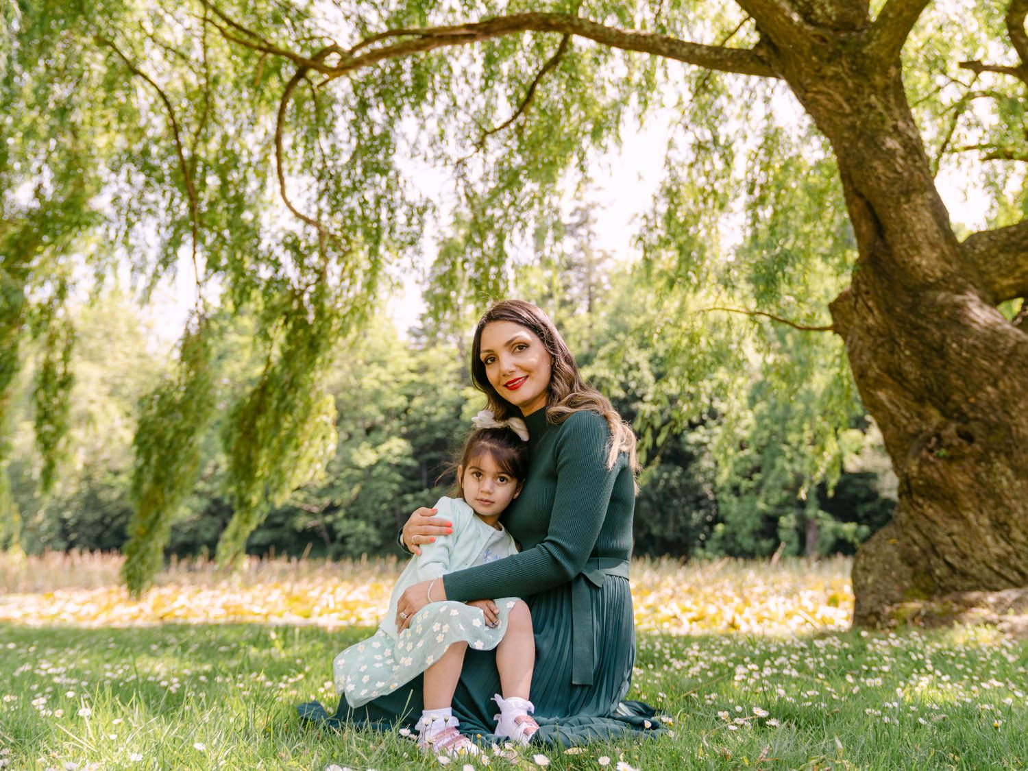 A mother and young daughter sitting together under a willow tree in a lush green park on a sunny day.