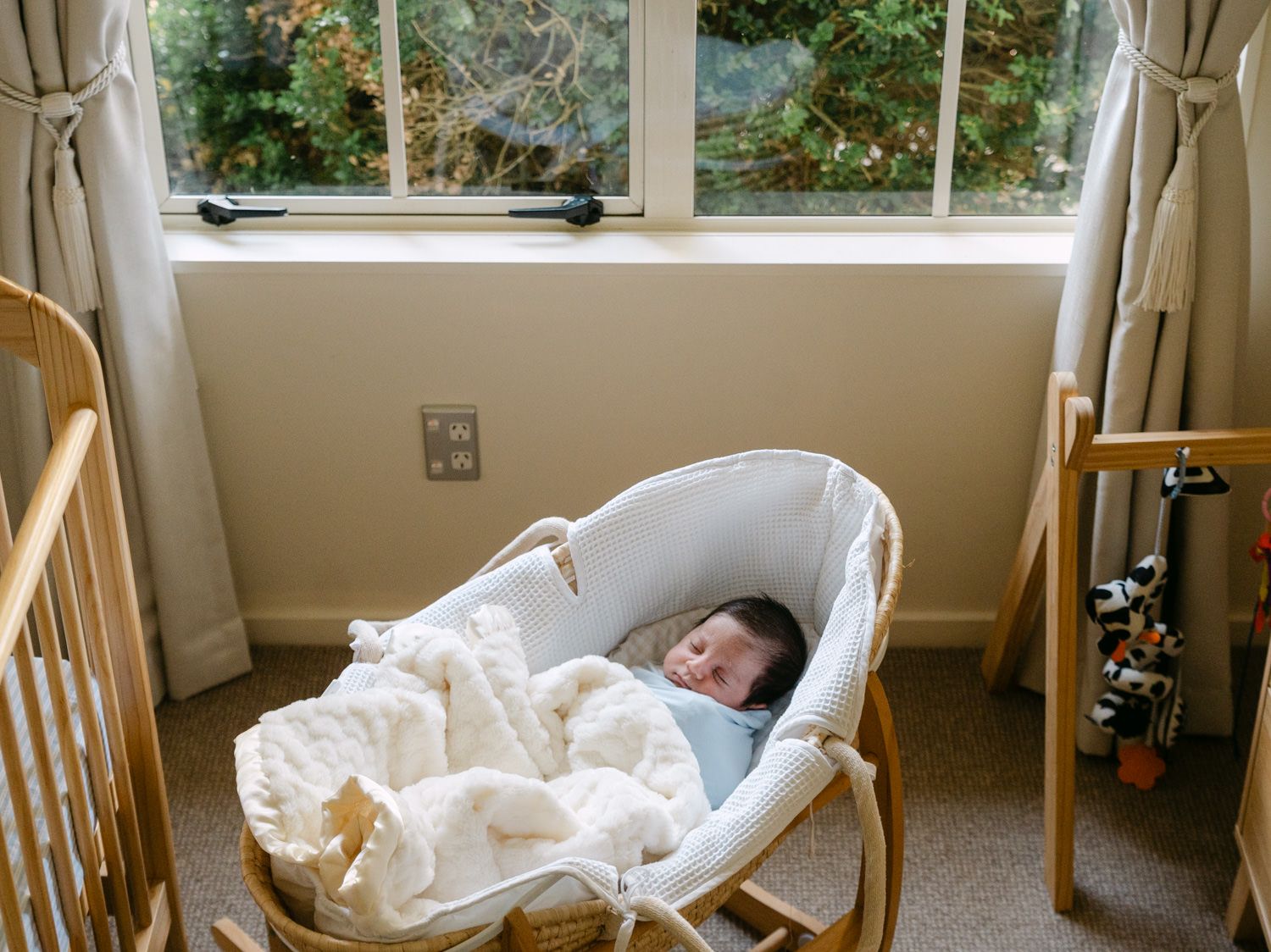 Sleeping newborn baby in a wicker Moses basket beside a wooden crib near a bright nursery window.