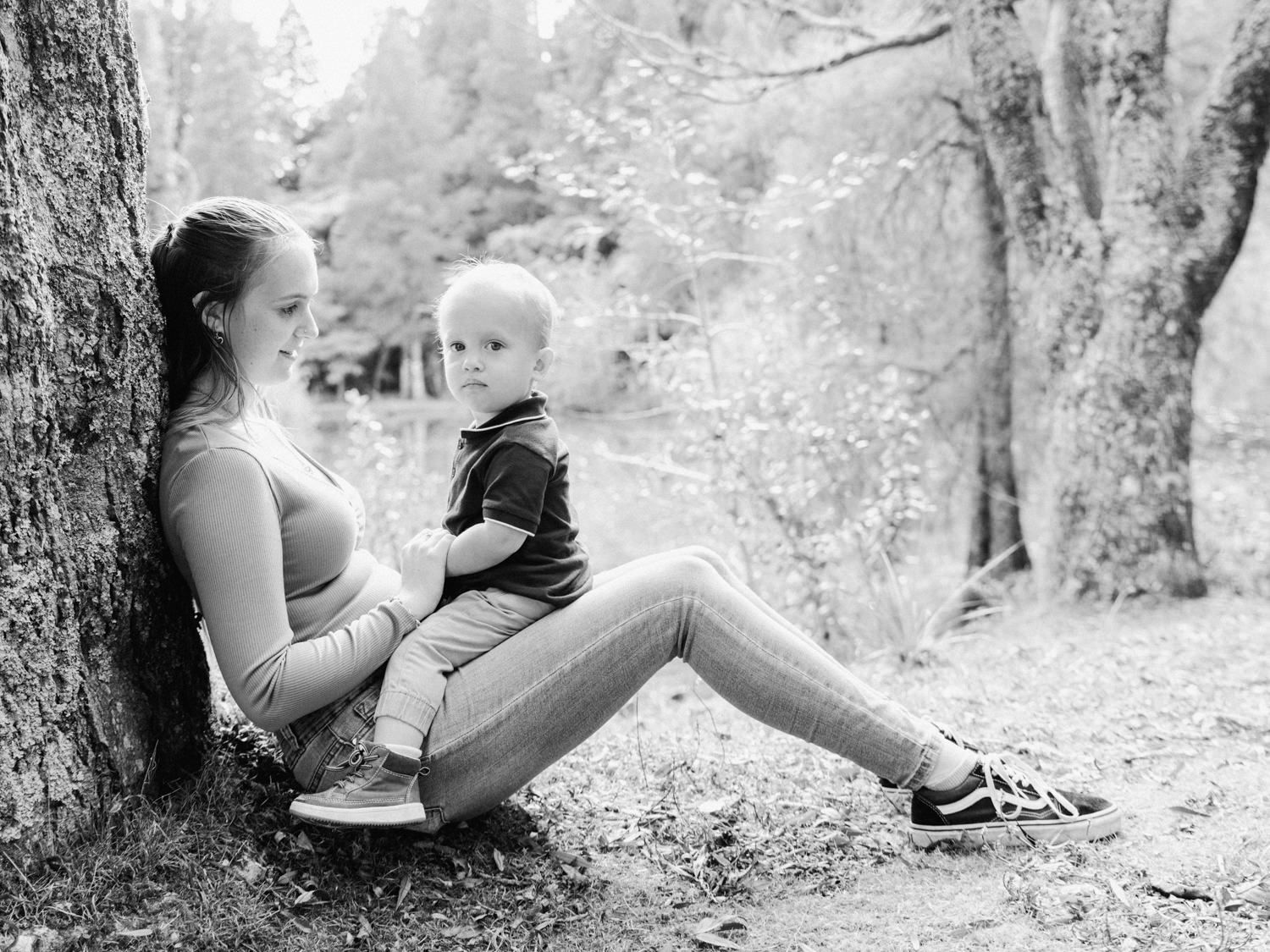 Black and white photo of a young mother sitting against a tree with her toddler son on her lap outdoors.