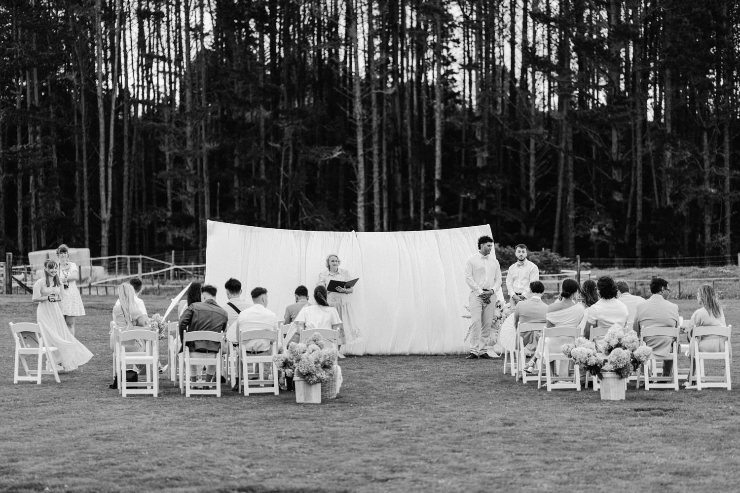 Black and white outdoor wedding ceremony with guests seated on white chairs facing altar backdrop surrounded by tall pine trees.
