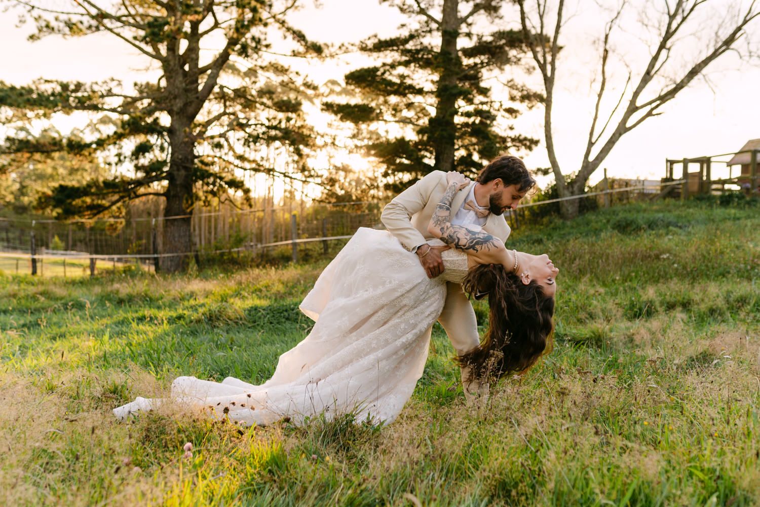 Couple sharing a romantic dip kiss in a sunlit grassy field at golden hour, bride in elegant white gown.