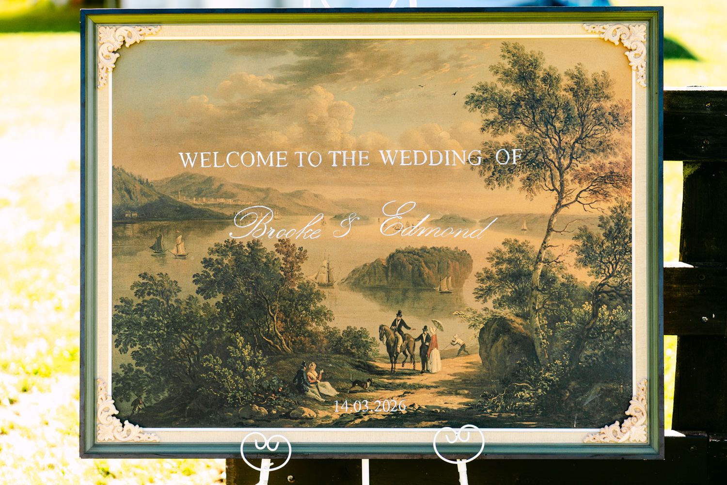 Elegant wedding welcome sign with a scenic landscape painting backdrop displayed outdoors.
